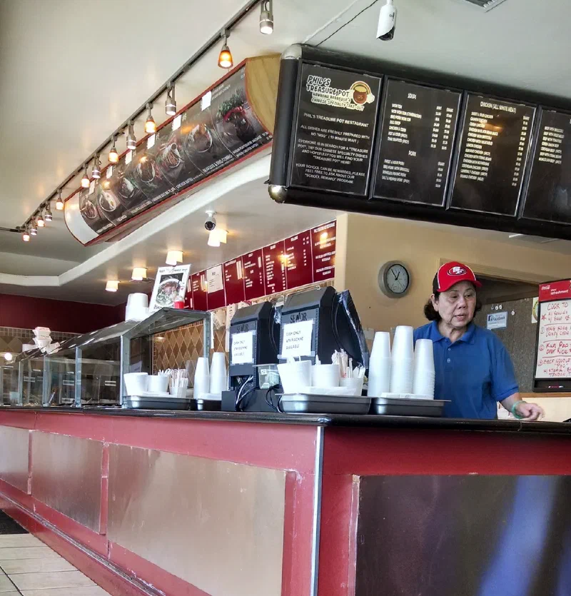 The order counter inside Phil’s: steam trays behind glass, stacks of takeout cups, and overhead menu boards.
