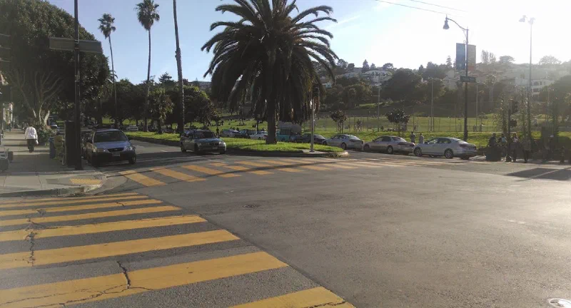 Late afternoon sun across Dolores Park — palm trees, a lawn dotted with people, and a wide empty crosswalk in the foreground.