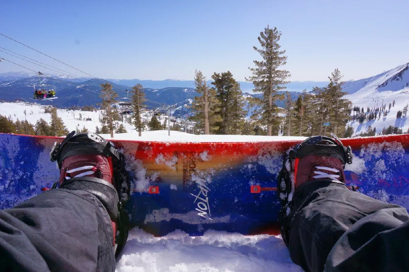 Looking down from a chairlift at a pair of boots strapped into a snowboard, with snow-covered pines and Lake Tahoe beyond.